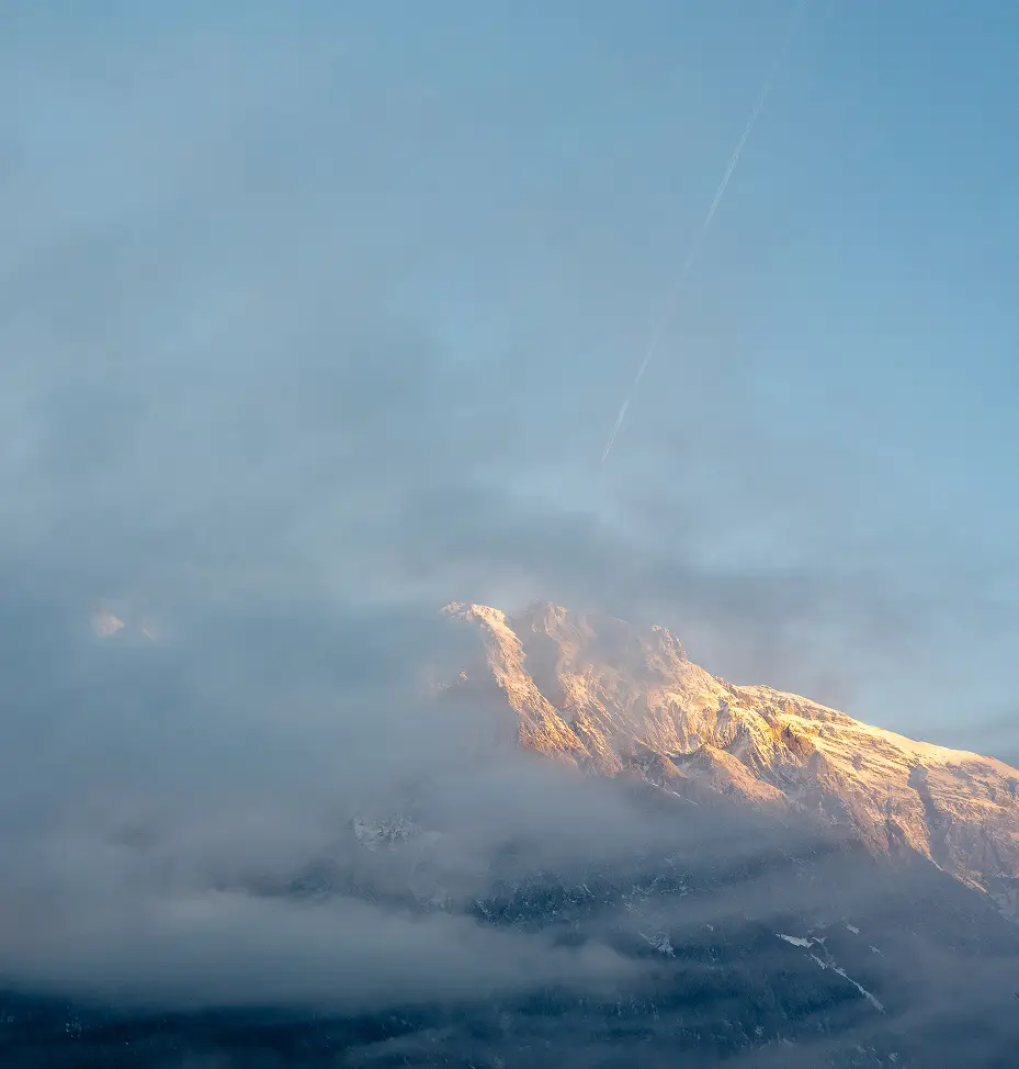 Majestic snow-covered mountain peak illuminated by the golden hour sun, partially hidden by soft clouds with a clear plane contrail in the blue sky.