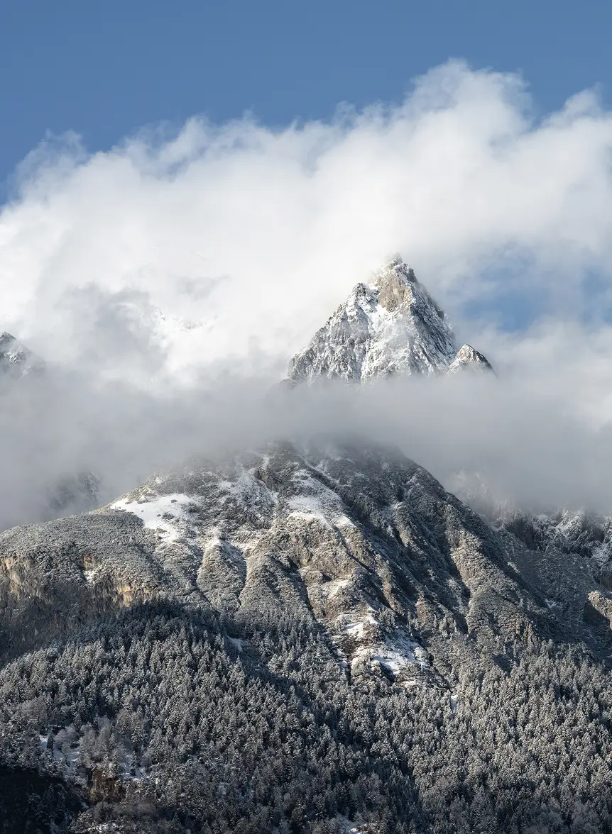 Detailed view of a rugged, snow-capped mountain peak emerging through a layer of thick white clouds under a clear sky