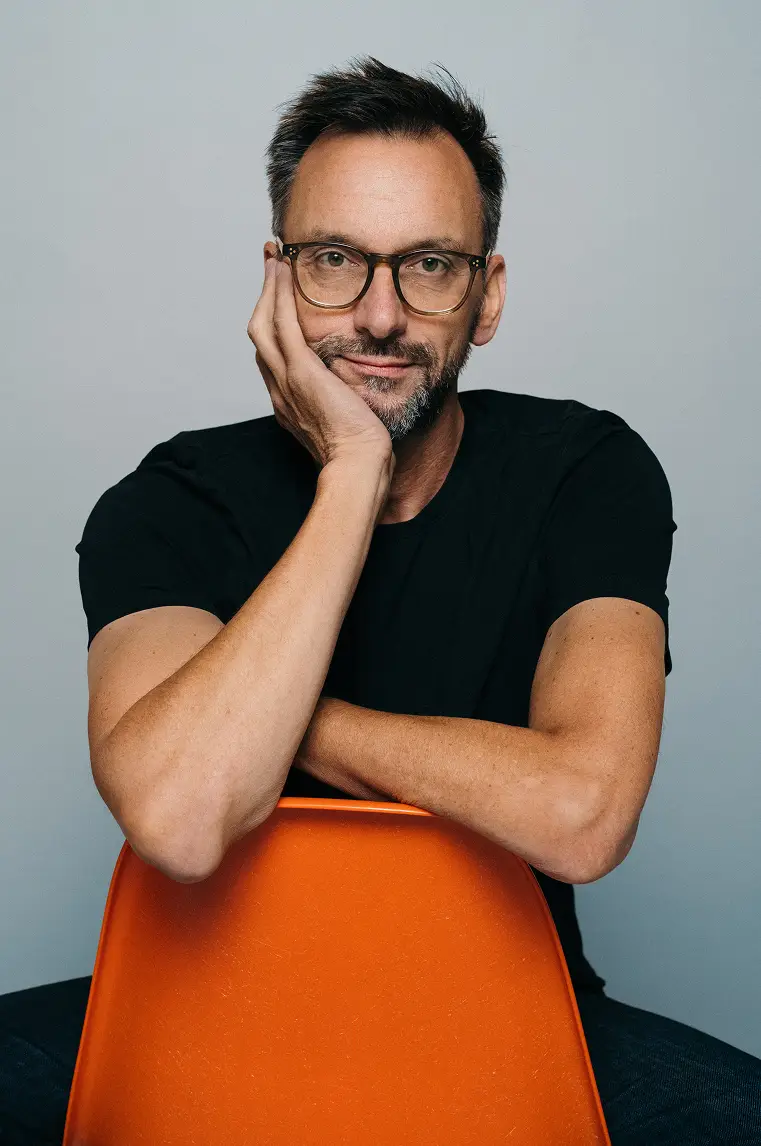 Martin Gundolf sitting cross-legged on a bright orange chair, wearing a black t-shirt and glasses against a neutral backgroun