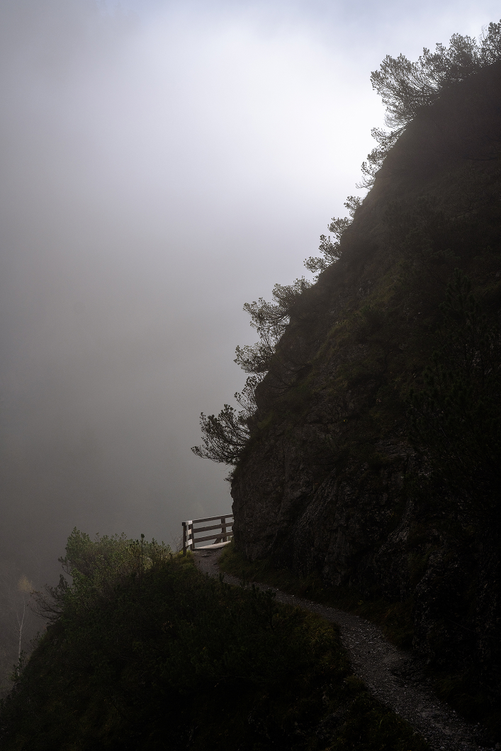 A narrow mountain path with a wooden railing next to a dark, steep cliff, disappearing into thick white fog